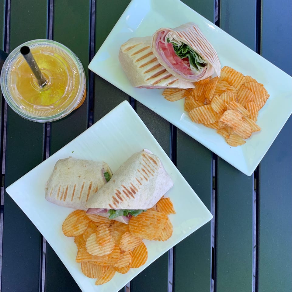 Two grilled wraps with chips on white plates and an orange drink with a straw on a green table.