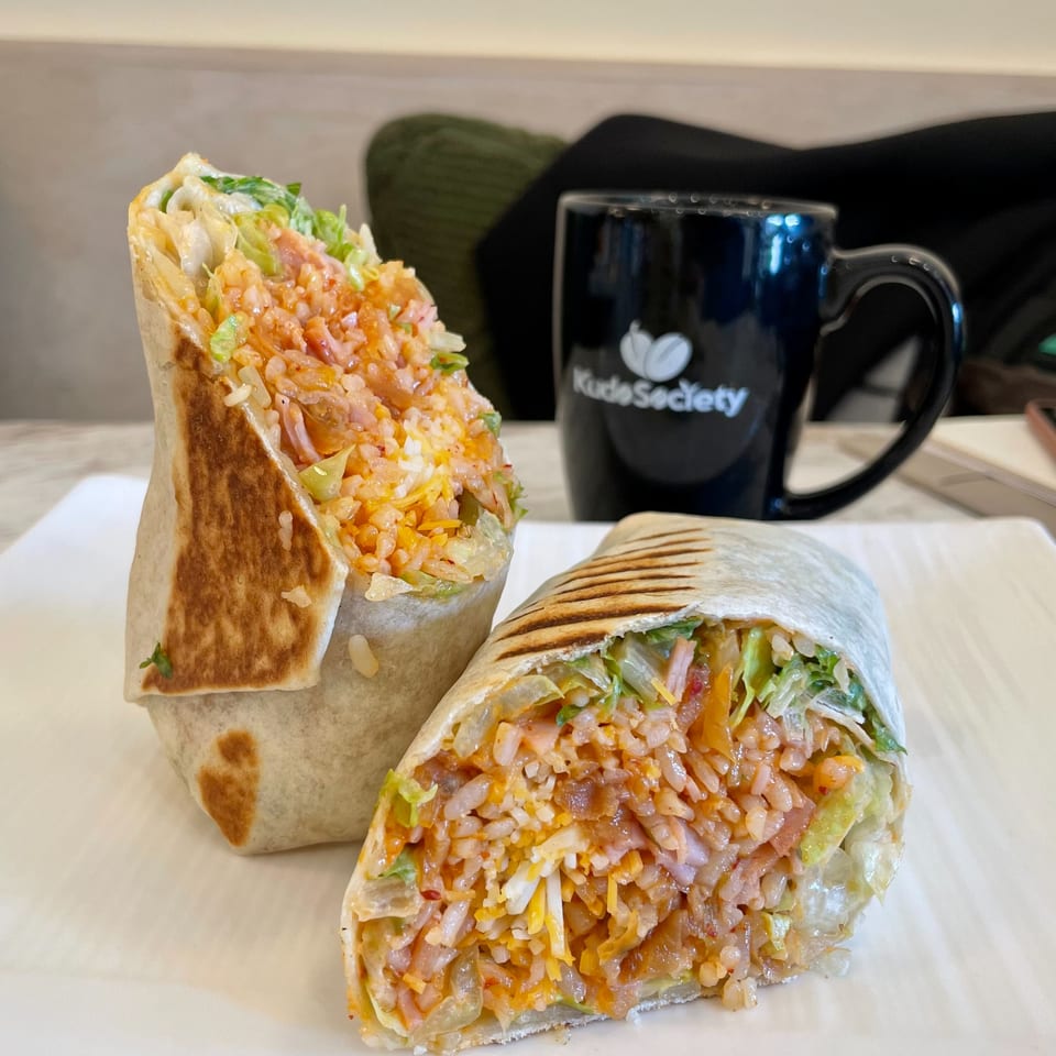 Two halves of a Kudo Society burrito filled with rice, meat, cheese, and lettuce, next to a branded mug.