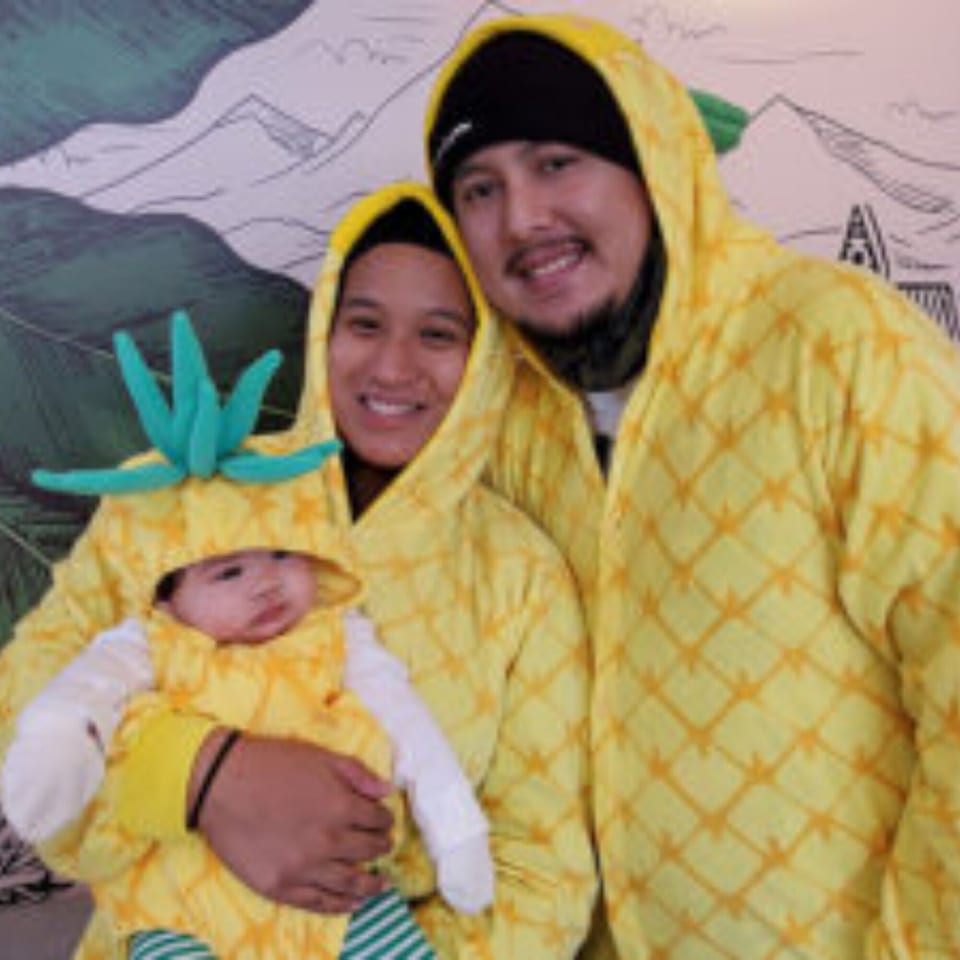 A family of three, including a baby, dressed in matching pineapple-themed outfits.