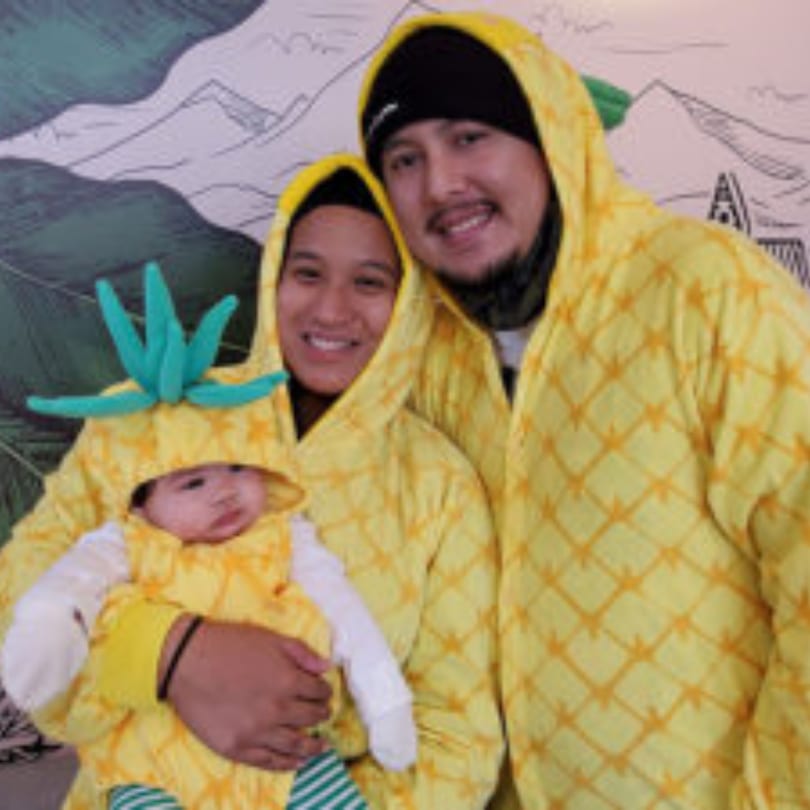 A family of three, including a baby, dressed in matching pineapple-themed outfits.