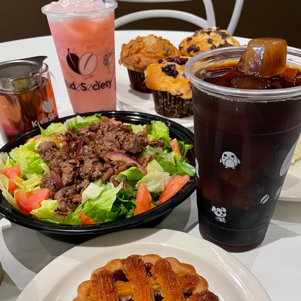Kudo Society's beef salad, muffins, iced coffee, pink drink, and a fruit tart on a white table.