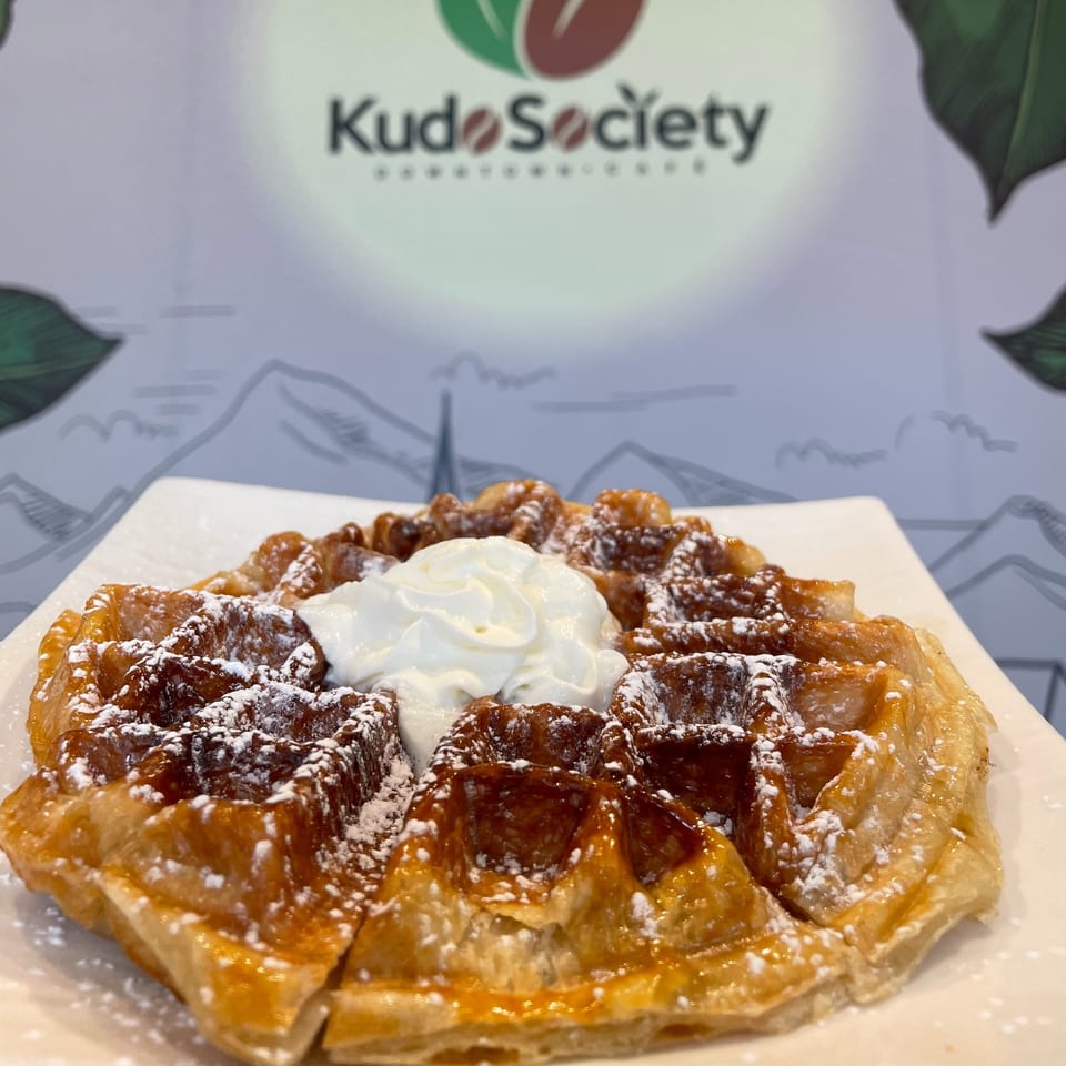 A golden Kudo Society waffle, topped with whipped cream and powdered sugar, on a white plate.