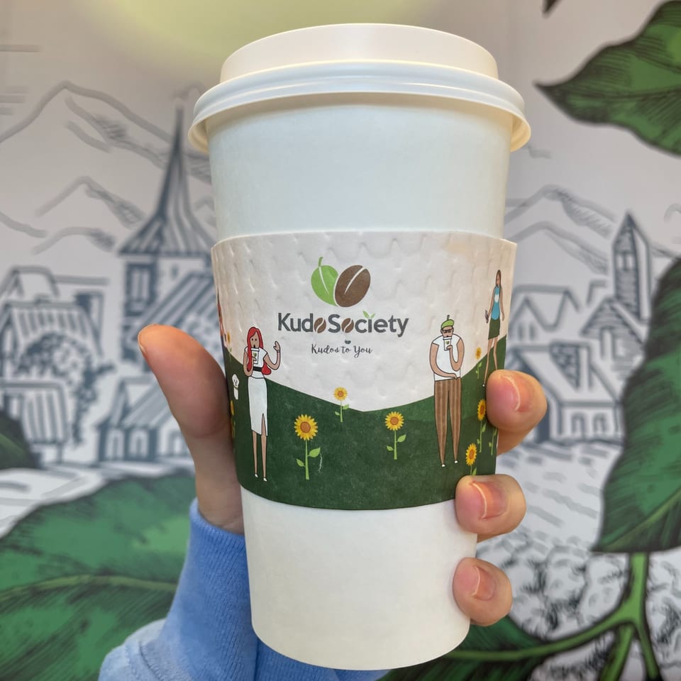 Hand holding a Kudo Society coffee cup with a custom sleeve featuring people and sunflowers.