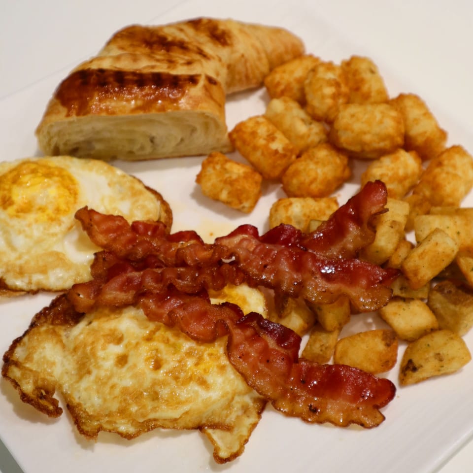 A breakfast plate with crispy bacon, fried eggs, golden tater tots, and a croissant.